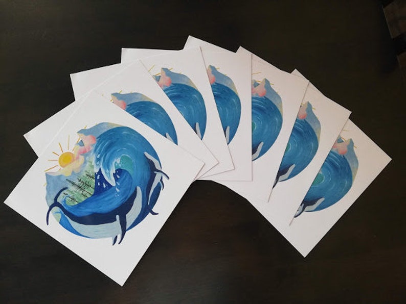 May include: A set of eight blank greeting cards featuring a watercolor illustration of a whale breaching in a wave with a sun and trees in the background. The cards are white with a blue and green watercolor design.