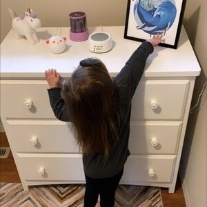 May include: A white dresser with three drawers and white knobs. A child is standing in front of the dresser, reaching up to touch a framed print of a whale in a wave. There are three small toys on top of the dresser, including a unicorn, a white noise machine, and a nightlight.