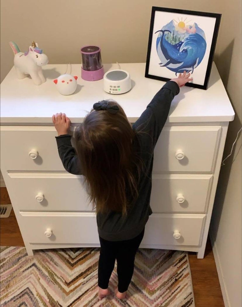 May include: A white dresser with three drawers and white knobs. A child is standing in front of the dresser, reaching up to touch a framed print of a whale in a wave. There are three small toys on top of the dresser, including a unicorn, a white noise machine, and a nightlight.