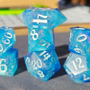 May include: Five blue and white iridescent dice with white numbers. The dice are stacked in a pyramid shape with the top die showing a 17, the next die down showing a 14, the next die down showing a 6, and the two bottom dice showing a 12 and a 6.