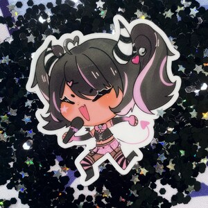 May include: A sticker featuring a cartoon character with black hair, pink highlights, and a black and white striped headband. The character is wearing a black and pink outfit and holding a microphone. The sticker is on a black and silver glitter background.