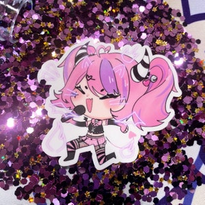 May include: A pink and purple cartoon character with black and white striped horns and a tail. The character is singing into a microphone and has a pink heart on its cheek. The character is on a background of purple and gold glitter.