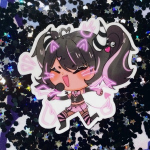 May include: A sticker featuring a cartoon character with black hair, pink highlights, and a pink and black outfit. The character is holding a microphone and has a pink heart on their cheek. The sticker has a pink and white outline and is set against a black and silver glitter background.