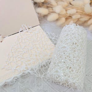 May include: A white, glittery, mesh ribbon rolled up on a white surface. The ribbon has a delicate, lacy pattern.