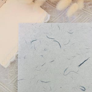 以下が含まれることがあります： 薄い灰色の紙のクローズアップ。青い繊維が全体に埋め込まれています。紙は質感があり、わずかに光沢があります。