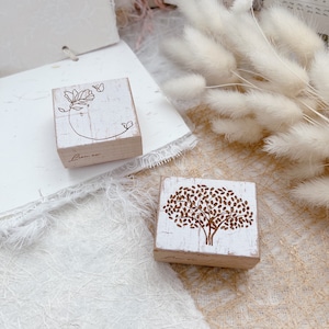 May include: Two wooden stamps with floral designs. The first stamp features a flower with a butterfly and the text "Bloom now". The second stamp features a tree with leaves.