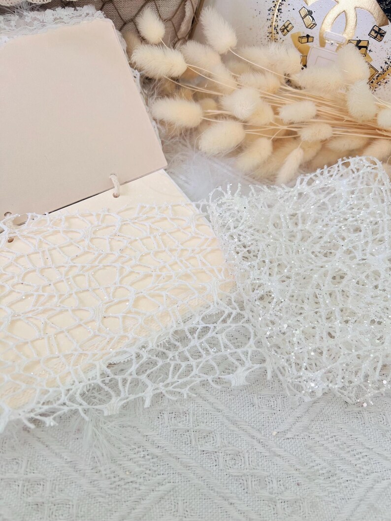 May include: Two pieces of white lace fabric with a delicate, intricate pattern. One piece is smooth and the other has a subtle sparkle.