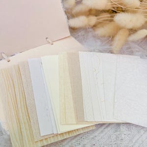 May include: A selection of textured paper sheets in cream, beige, and white. The papers are fanned out, with a soft pink notebook in the background. Dried fluffy beige flowers are also visible.