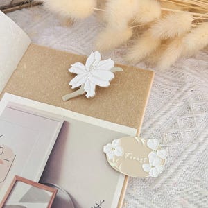 May include: Two white enamel pins with floral designs. One pin features a single flower with a stem and the word "Forest" in cursive. The other pin features a cluster of flowers.