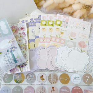 May include: A selection of floral-themed stationery, including stickers, decorative paper, and notecards. The stickers are round and feature various designs, including flowers and inspirational phrases. The colour palette includes pastels like pink, lavender, and green.