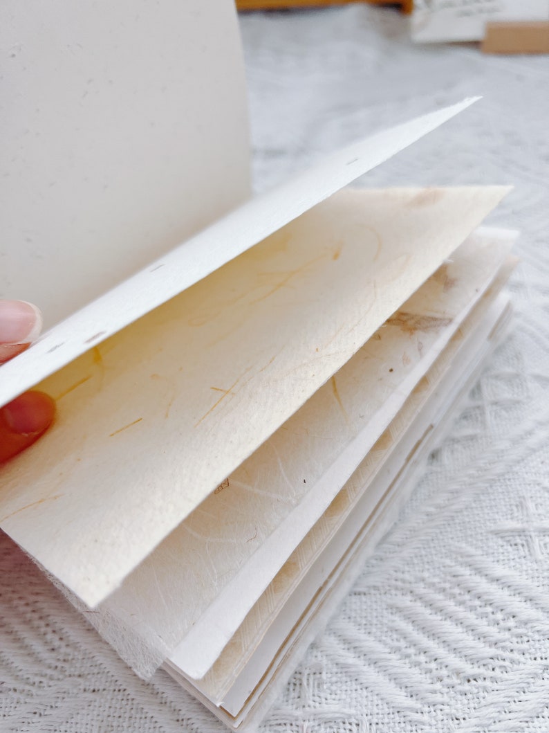 以下が含まれることがあります： クリーム色のページが積み重ねられた手作りノート。ページは、繊維が見える質感のある紙で作られています。
