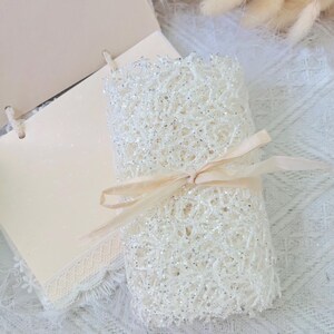 May include: A white lace fabric roll with a delicate, sparkly texture, tied with a beige ribbon. The fabric is rolled up and tied with a ribbon, creating a beautiful and elegant presentation.