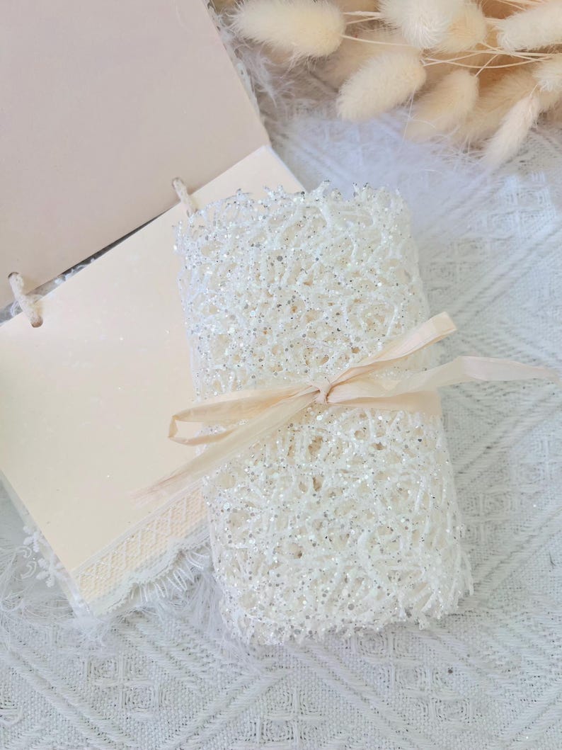 May include: A white lace fabric roll with a delicate, sparkly texture, tied with a beige ribbon. The fabric is rolled up and tied with a ribbon, creating a beautiful and elegant presentation.