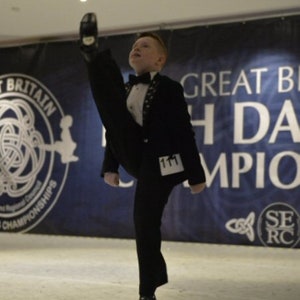 May include: A young boy in a black suit and a white bow tie performs a dance move with his leg extended and his foot pointed. He is wearing a number tag that says "111". The background is a blue banner with the text "Great Britain Irish Dancing Championships".