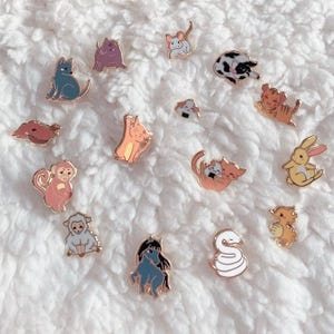 May include: A selection of enamel pins with animal designs, such as cats, a rabbit, and a snake. The pins have gold-coloured metal borders and are set against a white, fluffy background. The pins are in various colours.