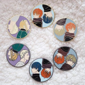 Op de afbeelding: Zes ronde emaille pins met gouden rand. Elke pin toont twee zijprofielen van anime-stijl personages in verschillende kleurencombinaties, waaronder rood, blauw, blond en paars. De achtergronden zijn lichtblauw en paars.