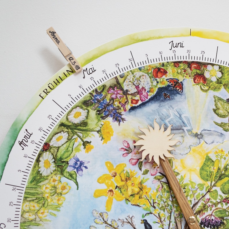 May include: A colorful, circular wall clock with a watercolor floral design. The clock face features months in German, a wooden sun-shaped pointer, and a wooden clip with the name "Anna" and the date "12.5.18". The clock is decorated with various flowers and butterflies.