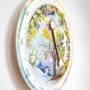 May include: A round, wooden wall clock featuring a watercolor illustration of the four seasons. The clock face displays a tree at the center, surrounded by seasonal imagery and a wooden sun-shaped hand. The clock is mounted on a white wall.