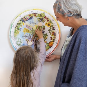 May include: A colorful, circular educational calendar featuring illustrations of the seasons. A child and an adult interact with the calendar, pointing to different sections. The calendar is decorated with flowers, fruits, and seasonal imagery.