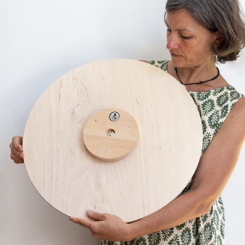 May include: A large, round wooden board with a natural finish. A smaller, circular wooden piece is attached to the center with screws, featuring a metal component. The board is being held by a person.