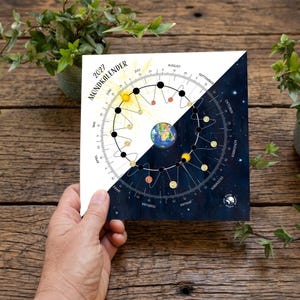Könnte beinhalten: Ein Mondkalender für 2027 mit einem kreisförmigen Diagramm der Erde und der Mondphasen. Das Design ist diagonal geteilt zwischen einem weißen Hintergrund mit einer Sonnendarstellung und einem dunkelblauen Sternenhimmel.