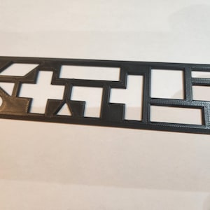 May include: A black rectangular plastic stencil featuring various geometric cutouts, including a circle, a triangle, a cross, and several rectangles of different sizes, all arranged within a flat, grid-like frame with a textured surface finish.