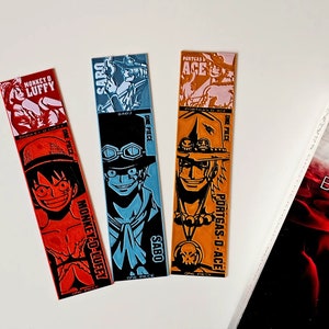 May include: Three colorful bookmarks featuring characters from the anime One Piece. The bookmarks feature Luffy, Sabo, and Ace. Each bookmark has the character's name and the words "One Piece" printed on them.