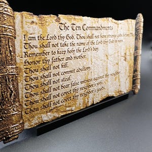 May include: A decorative scroll featuring the Ten Commandments. The text is inscribed on a textured, aged-looking surface with a brown and gold border. The scroll is displayed on a black stand, suitable for home decor or religious settings.