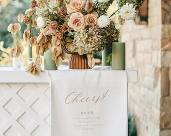 May include: A floral arrangement in a ribbed brown bowl featuring white dahlias, peach roses, and dried foliage, flanked by two sage green ribbed pillar candles on a white bar with a hanging menu banner that reads Cheers! Beer and Wine.