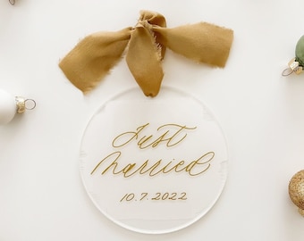 May include: A circular clear acrylic ornament featuring the gold script text Just Married and the date 10.7.2022. The ornament is topped with a mustard yellow frayed ribbon and is surrounded by various small gold, white, and sage green baubles.