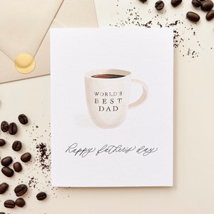 May include: A white card with a hand-drawn illustration of a coffee mug with the text "World's Best Dad" and the words "Happy Father's Day" written in cursive.