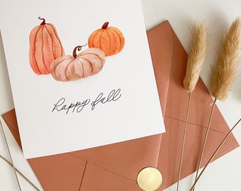 May include: A white card with a watercolor illustration of three pumpkins and the text "Happy Fall". The card is sitting on a white surface with a brown envelope and dried grass.