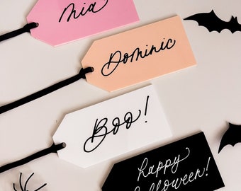 May include: Four Halloween-themed gift tags with black string. The tags are pink, peach, white, and black. The pink tag says "Mia", the peach tag says "Dominic", the white tag says "Boo!" and the black tag says "Happy Halloween!"