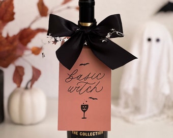 May include: A bottle of red wine with a black bow and a gift tag that says "Basic Witch". The tag has a black and white illustration of a wine glass with a jack-o-lantern face.