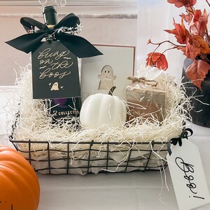 May include: A black wire basket filled with white shredded paper, a bottle of wine with a black bow and a tag that says "Boo! on the Boo's", a white ceramic ghost, a brown candle, and a white tag that says "Boo!"