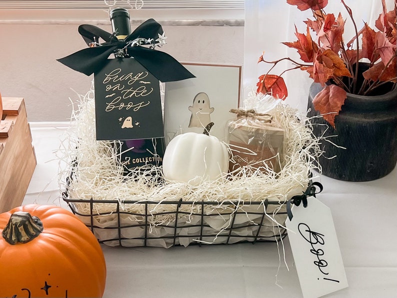 May include: A black wire basket filled with white shredded paper, a bottle of wine with a black bow and a tag that says "Boo! on the Boo's", a white ceramic ghost, a brown candle, and a white tag that says "Boo!"