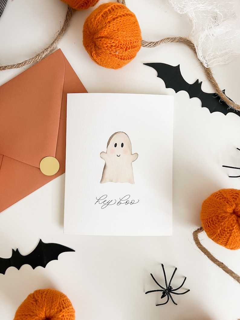 May include: A white card with a watercolour illustration of a ghost with a smiling face. The card says "Hey Boo" in black cursive writing. The card is surrounded by orange yarn pumpkins, black bats, and black spiders on a white background.