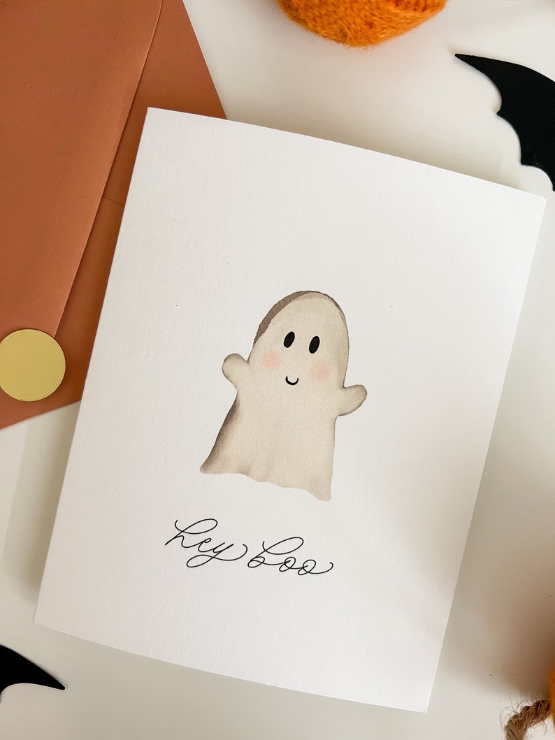 May include: A white card with a hand-drawn illustration of a ghost with a smiling face. The text "Hey Boo" is written in black cursive below the ghost.