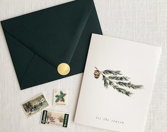 May include: A green envelope with a gold sticker and three Christmas stamps. A white card with a green pine branch and the text "tis the season" is on top of the envelope.