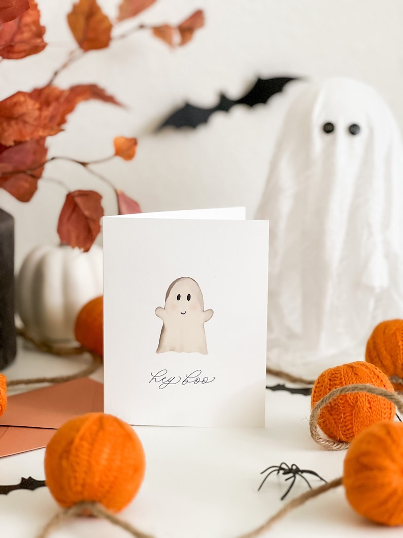 May include: A white card with a hand-drawn ghost and the text "Hey Boo" in black ink. The card is surrounded by orange yarn balls and a black spider.
