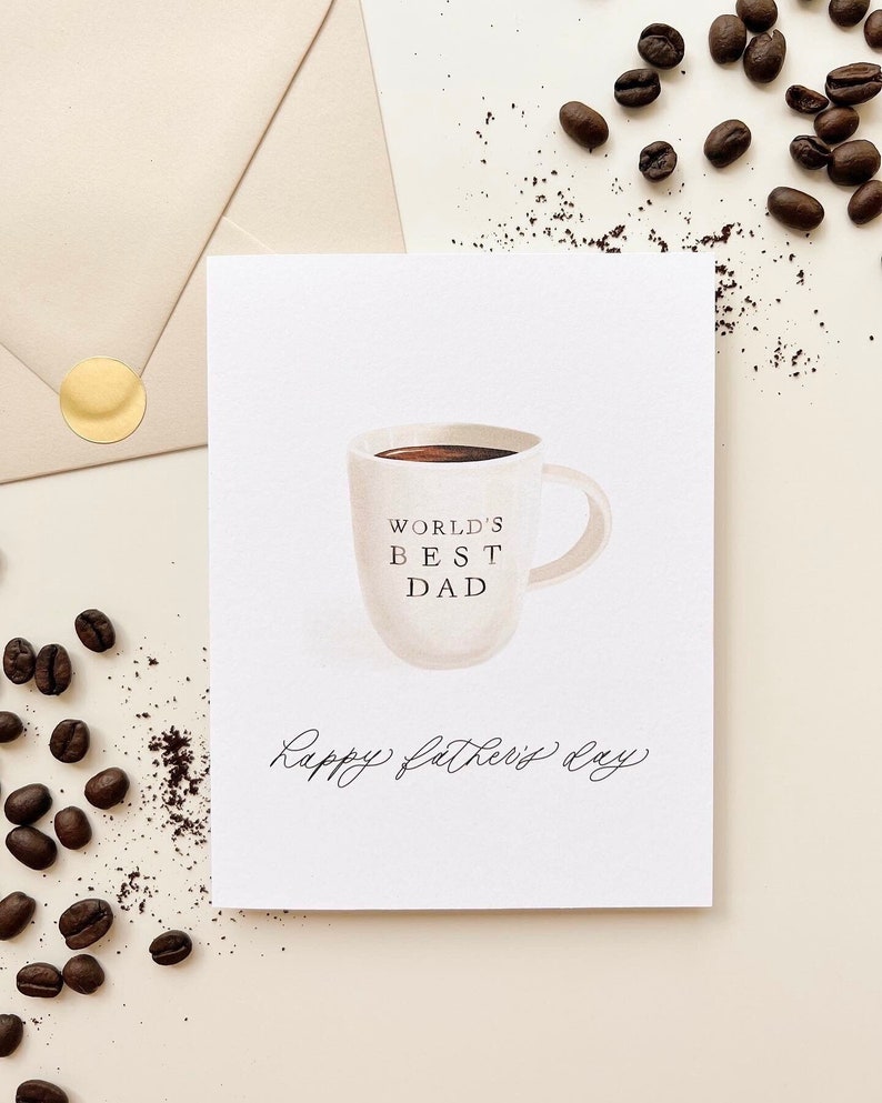 May include: A white card with a hand-drawn illustration of a coffee mug with the text "World's Best Dad" and the words "Happy Father's Day" written in cursive.