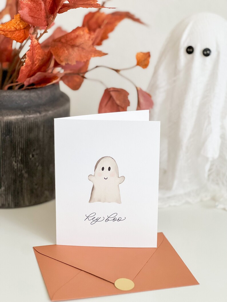 May include: A white card with a cute cartoon ghost and the text "Hey Boo" on it. The card is sitting on a peach-coloured envelope with a gold circle sticker.