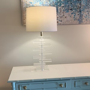 May include: A table lamp featuring a white drum shade and a clear acrylic base composed of stacked rectangular blocks. The lamp has a central metal rod supporting the shade and sits on a square acrylic plate, emitting a soft light from within.