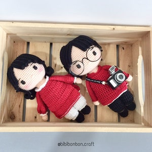 May include: Two crocheted dolls in a wooden crate. The dolls are wearing red sweaters and have black hair. One doll is holding a camera.