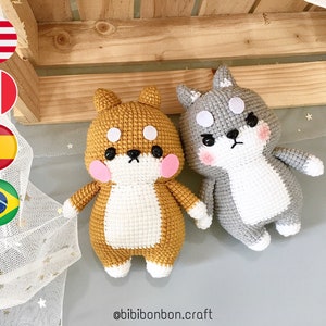 May include: Two crocheted stuffed dog toys. One is brown and white and the other is gray and white. Both have black eyes and pink cheeks. The toys are sitting on a gray surface with a white mesh fabric in the background.