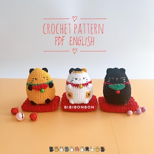 May include: Three crocheted Maneki-Neko, or lucky cat, figurines. The cats are sitting on red cushions and are wearing a red collar with a bell. The cats are yellow, white, and black. The image includes the text "CROCHET PATTERN PDF ENGLISH" and "BIBIBONBON".