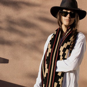 Może przedstawiać: Kobieta w białej koszuli i brązowym kapeluszu z czarno-brązowym wzorzystym szalikiem. Szalik ma czerwone paski.