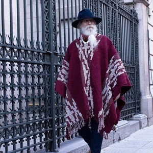 Puede incluir: Una persona con un poncho burdeos con un patr&oacute;n geom&eacute;trico blanco, un sombrero negro y vaqueros azules. El poncho tiene un dobladillo con flecos. La persona camina por una acera junto a una verja de hierro forjado.