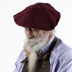 May include: A burgundy beret with a slightly distressed appearance. The beret is made of a soft fabric and has a classic, round shape. The hat is worn by a person with a long white beard and a blue striped shirt.
