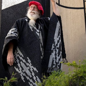 May include: A man with a long white beard and a red beret wears a black and white patterned poncho. He is standing in front of a wall with a black and white geometric design.